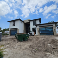 Miami construction site. Dirt and construction supplies sit in the foreground and a house under construction is in the background. The house is white with dark brown trim. The sky is partially cloudy.